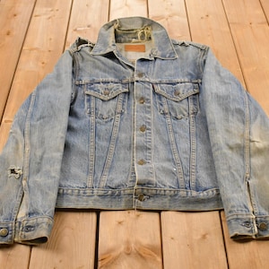 Könnte beinhalten: Eine hellblaue Jeansjacke mit klassischem Kragen, langen Ärmeln mit geknöpften Manschetten und zwei Brusttaschen mit Knopfklappen. Die Jacke weist ein abgenutztes Finish mit sichtbaren Gebrauchsspuren auf, einschließlich ausgefranster Kanten.