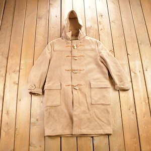 May include: A beige hooded duffle coat with brown leather toggles and loops. The coat has two large pockets on the front and is laid flat on a wooden surface.