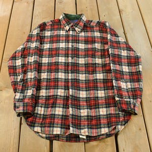 May include: A long-sleeved, button-down flannel shirt in a classic plaid pattern. The shirt features a red, black, white, and cream color scheme. It has a green collar lining and buttoned cuffs, suitable for casual wear.