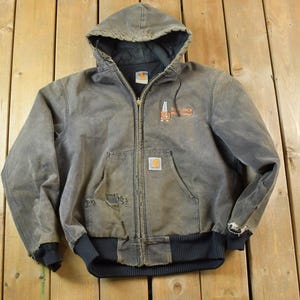 May include: A dark gray hooded jacket with a zipper closure and a Carhartt logo patch on the left chest. The jacket has a worn and distressed look, with visible wear and tear on the fabric.
