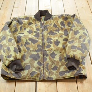 May include: A vintage camouflage bomber jacket with a full zipper closure. The jacket features a pattern of brown and olive green shapes on a tan background. It has dark brown ribbed cuffs and collar.