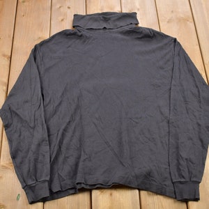 Pode incluir: Uma camisa de gola alta de manga comprida, cinza escura. A camisa é feita de um tecido macio e confortável e tem um caimento relaxado.