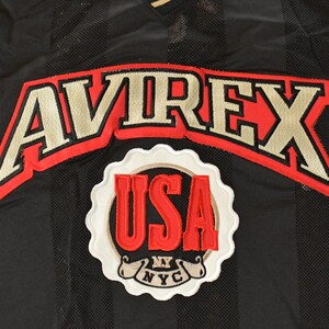 Op de afbeelding: Zwarte mesh stof met verticale strepen, met de geborduurde tekst "AVIREX" in goud met een rode omlijning. Daaronder toont een witte, geschulpte patch "USA" in rood, met "NY NYC" eronder.