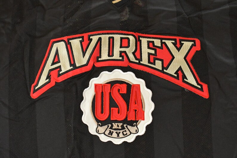 Op de afbeelding: Zwarte mesh stof met verticale strepen, met de geborduurde tekst "AVIREX" in goud met een rode omlijning. Daaronder toont een witte, geschulpte patch "USA" in rood, met "NY NYC" eronder.