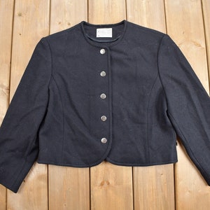 K&ouml;nnte beinhalten: Schwarze, kurze Jacke mit rundem Ausschnitt und langen &Auml;rmeln. Die Jacke hat eine Reihe von f&uuml;nf silbernen Kn&ouml;pfen auf der Vorderseite. Das Kleidungsst&uuml;ck ist aus einem dunklen Stoff gefertigt und wird auf einer hellen Holzoberfl&auml;che pr&auml;sentiert.