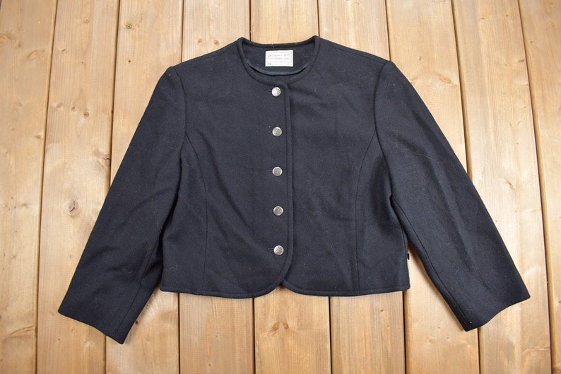 K&ouml;nnte beinhalten: Schwarze, kurze Jacke mit rundem Ausschnitt und langen &Auml;rmeln. Die Jacke hat eine Reihe von f&uuml;nf silbernen Kn&ouml;pfen auf der Vorderseite. Das Kleidungsst&uuml;ck ist aus einem dunklen Stoff gefertigt und wird auf einer hellen Holzoberfl&auml;che pr&auml;sentiert.