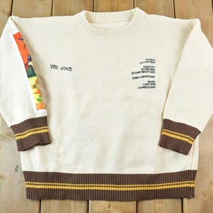 Puede incluir: Un suéter de color crema con rayas marrones y amarillas en los puños y el dobladillo inferior. El suéter tiene un gráfico colorido en la manga izquierda y texto que dice "YOU GOT IT" y "ROAD EXTENDED LA VERANDA HARVEST SCENE HIGH ST. MARY'S STREET SUNFLOWER CAFE ROAD COMMUNITY CENTER".
