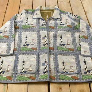 May include: A light blue jacket featuring a repeating design of lighthouses, sailboats, and coastal landscapes. The jacket has a button-up front, two pockets, and a grey trim. The fabric looks like a woven tapestry.