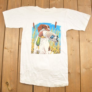 May include: White t-shirt with a graphic print of a brown and white dog carrying a duck in its mouth. The dog is standing in a field of tall grass with blue sky in the background.