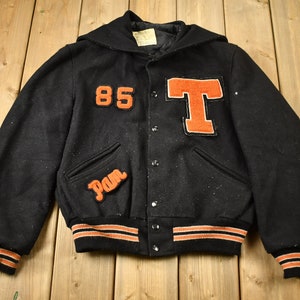 May include: Black varsity jacket with orange and white striped cuffs and collar. The jacket has a large orange "T" and the number "85" on the front. The word "Pam" is embroidered on the left pocket.