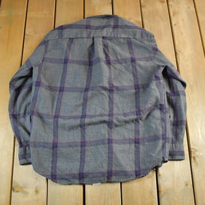 Puede incluir: Camisa a cuadros gris y morado. La camisa est&aacute; hecha de una tela suave y presenta un patr&oacute;n de cuadros cl&aacute;sico en gris y morado. Tiene mangas largas y una botonadura.