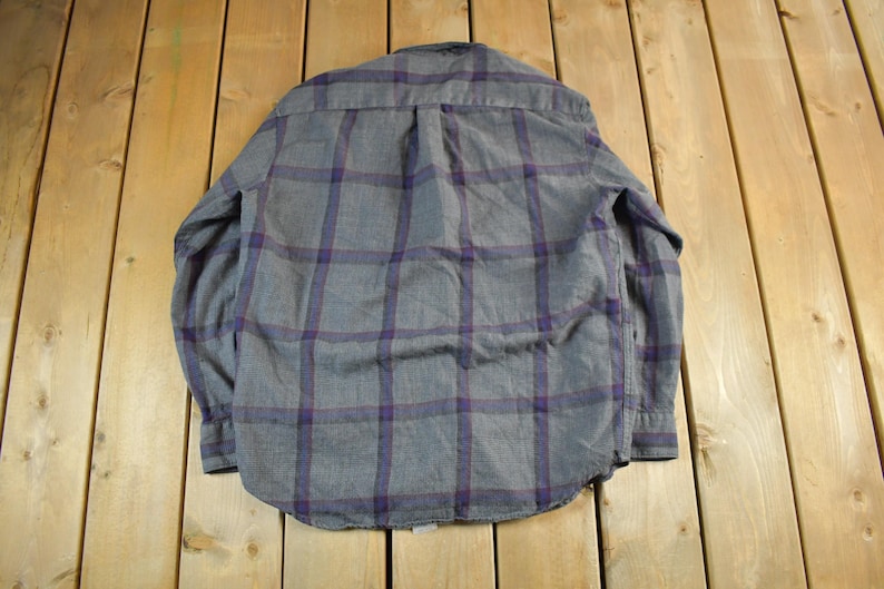 Puede incluir: Camisa a cuadros gris y morado. La camisa est&aacute; hecha de una tela suave y presenta un patr&oacute;n de cuadros cl&aacute;sico en gris y morado. Tiene mangas largas y una botonadura.