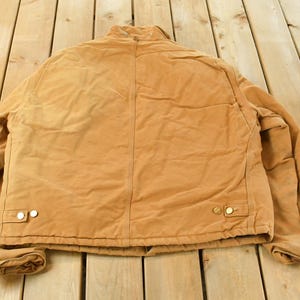 Puede incluir: Una chaqueta de trabajo de color marr&oacute;n con cierre de botones y dos bolsillos delanteros. La chaqueta est&aacute; hecha de un material duradero y tiene un dise&ntilde;o cl&aacute;sico. La chaqueta est&aacute; sobre una superficie de madera.