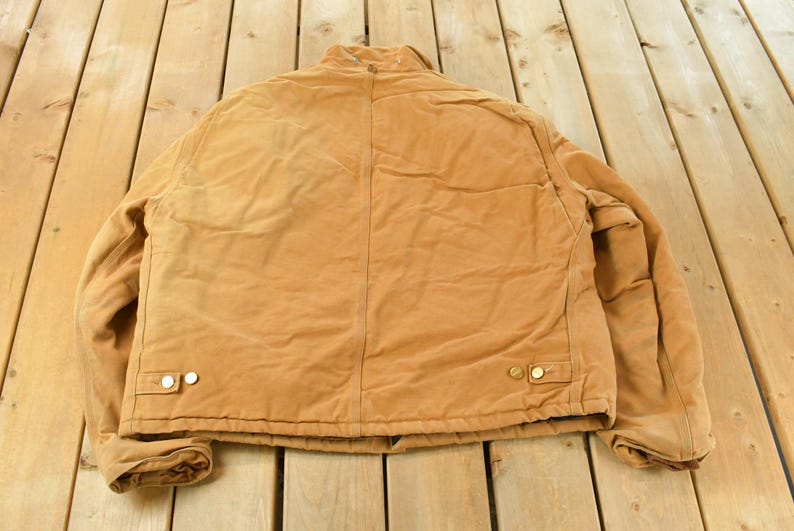 Puede incluir: Una chaqueta de trabajo de color marr&oacute;n con cierre de botones y dos bolsillos delanteros. La chaqueta est&aacute; hecha de un material duradero y tiene un dise&ntilde;o cl&aacute;sico. La chaqueta est&aacute; sobre una superficie de madera.