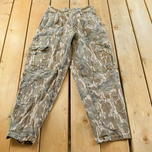 May include: Camouflage cargo pants in shades of brown, green, and gray, featuring multiple pockets and a button and zipper closure. The pants are laid flat on a wooden surface.