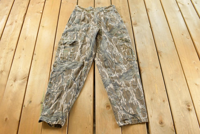 May include: Camouflage cargo pants in shades of brown, green, and gray, featuring multiple pockets and a button and zipper closure. The pants are laid flat on a wooden surface.