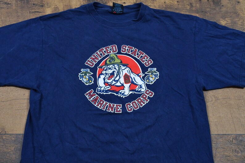 May include: A navy blue t-shirt with a graphic of the United States Marine Corps logo. The logo features a bulldog wearing a military hat and holding a rifle. The words "United States Marine Corps" are written around the logo.