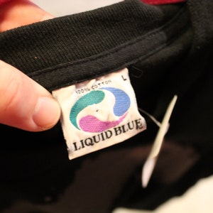 May include: A black Liquid Blue t-shirt tag with white text and a blue, green, and purple logo. The tag says "100% cotton" and "Liquid Blue" and has a size "L" label.