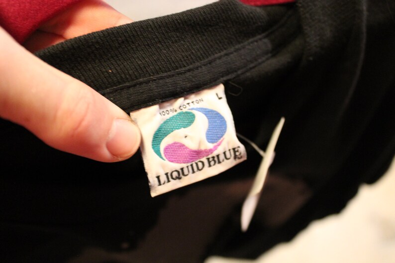 May include: A black Liquid Blue t-shirt tag with white text and a blue, green, and purple logo. The tag says "100% cotton" and "Liquid Blue" and has a size "L" label.