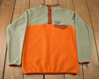 May include: A green and orange fleece pullover with a snap button placket and a brown patch pocket. The Patagonia logo is visible on the chest.