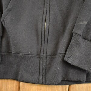 May include: Close-up of a black zip-up hooded sweatshirt. The cuffs and waistband are ribbed. The zipper is centered, and the fabric appears to be a soft, worn material. The sweatshirt is a casual garment.