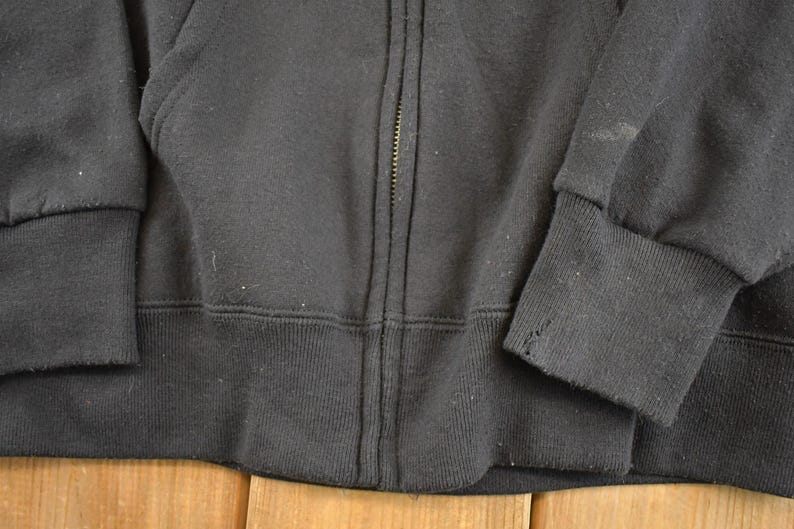 May include: Close-up of a black zip-up hooded sweatshirt. The cuffs and waistband are ribbed. The zipper is centered, and the fabric appears to be a soft, worn material. The sweatshirt is a casual garment.
