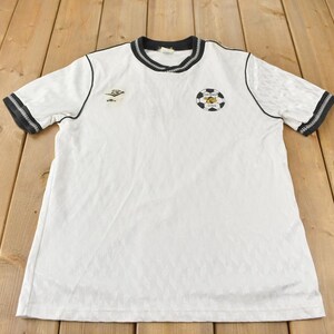 May include: White short-sleeved t-shirt with black trim around the collar and sleeves. The shirt has two embroidered patches, one with a soccer ball and the other with a logo.