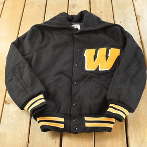 以下が含まれることがあります： 胸元に大きな黄色い「W」の文字のワッペンが付いた黒のバーシティジャケット。幅広の襟、フロントのスナップボタン留め、黄色と黒のストライプが入ったリブ編みの袖口と裾が特徴です。厚手の生地で作られています。