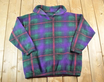 Puede incluir: Un jersey de forro polar vintage con un diseño de cuadros. Los colores son morado, verde y rojo. El jersey tiene cuello y cremallera de un cuarto. Las mangas son largas y el dobladillo es elástico. La tela parece suave y cálida.