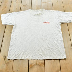 Op de afbeelding: Een wit t-shirt met korte mouwen en het oranje STIHL-logo op de linkerborst. Het kledingstuk heeft een ronde hals en een standaard pasvorm, gemaakt van een lichtgewicht stof met zichtbare natuurlijke plooien.