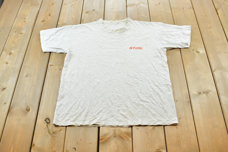 Op de afbeelding: Een wit t-shirt met korte mouwen en het oranje STIHL-logo op de linkerborst. Het kledingstuk heeft een ronde hals en een standaard pasvorm, gemaakt van een lichtgewicht stof met zichtbare natuurlijke plooien.