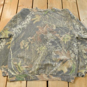 May include: A camouflage-patterned sweatshirt in shades of green, brown, and gray, featuring a crew neck and long sleeves. The fabric displays a detailed woodland design, ideal for outdoor activities or casual wear.