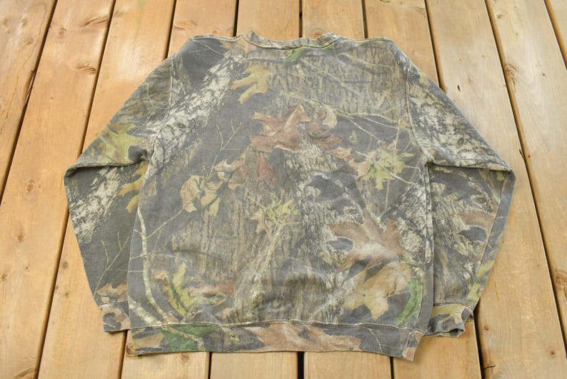 May include: A camouflage-patterned sweatshirt in shades of green, brown, and gray, featuring a crew neck and long sleeves. The fabric displays a detailed woodland design, ideal for outdoor activities or casual wear.