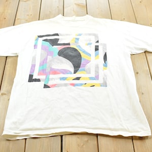 May include: A white short-sleeve t-shirt featuring a central graphic design composed of overlapping squares and abstract shapes in shades of black, yellow, light blue, and purple. The shirt has a crew neckline and a standard straight hemline.
