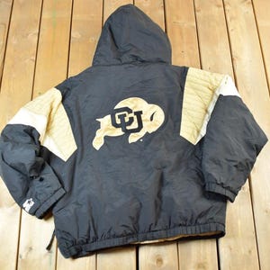 May include: Black and gold hooded jacket featuring the CU Buffs logo on the back. The jacket has gold and white accents on the shoulders and sleeves. Made by Starter.