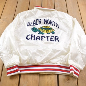 Puede incluir: Chaqueta de satén blanco con puños y cuello a rayas rojas y azules. La parte trasera presenta el texto "BLACK NORTH CHAPTER" sobre un gráfico de tortuga verde y amarilla. La chaqueta está sobre una superficie de madera.