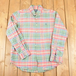 May include: A button-down plaid shirt in pastel colors of green, pink, and blue. The long-sleeved shirt has a button-down collar and a small embroidered logo on the chest. The shirt is laid flat on a wooden surface.