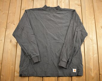 Pode incluir: Uma camisa de manga comprida cinza escura com gola alta e um pequeno patch com o logotipo da Carhartt na parte inferior direita.