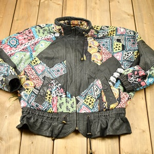 May include: A vintage black leather jacket with colourful patchwork panels. The jacket features a high collar, a zip-up front, and elastic cuffs and waist. The patchwork includes various geometric and floral patterns in shades of pink, blue, yellow, and green.