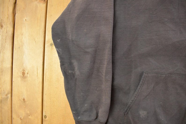 May include: A dark gray hoodie with a front pocket and a drawstring hood. The sleeve is visible, showing some discoloration. The background is a light brown wooden surface.