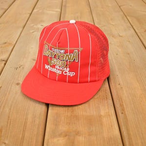 May include: A red Daytona 500 Winston Cup cap with white vertical stripes and a mesh back. The cap features the text "1993 Daytona 500 NASCAR Winston Cup" in gold and white lettering. The cap is on a wooden surface.