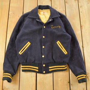 Peut inclure: Veste de style universitaire bleu marine vintage avec des détails jaunes. La veste présente une doublure jaune, le nom "Terry" brodé en jaune et des rayures jaunes sur les poignets et la taille. Elle a deux poches avant avec une bordure jaune.