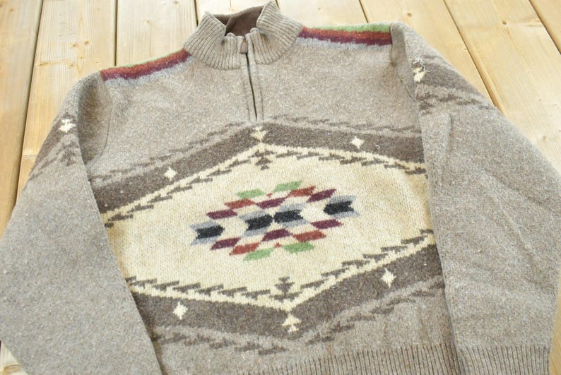 Peut inclure: Un pull gris zipp&eacute; avec un motif g&eacute;om&eacute;trique au centre. Le motif comprend des losanges cr&egrave;me, marron, noirs, rouges et verts. Le pull a un ourlet et des poignets c&ocirc;tel&eacute;s.