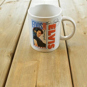 Puede incluir: Una taza de cerámica blanca con un gráfico de Elvis Presley, el texto Elvis Always the original y una firma. El diseño incluye un borde azul y rojo, un retrato de Elvis y el nombre Elvis impreso en letras grandes de color naranja.