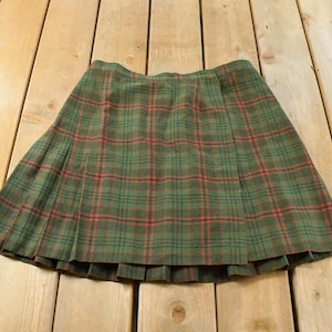 Puede incluir: Una falda hasta la rodilla con un patrón de cuadros escoceses en verde, negro y rojo. La prenda está diseñada con un panel frontal estilo cruzado y una serie de pliegues verticales que se extienden por la parte inferior de la falda.
