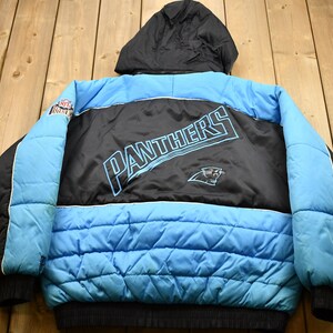 Op de afbeelding: Een zwart en blauw pufferjack met een Carolina Panthers logo op de rug. Het jack heeft een capuchon en een ritssluiting.