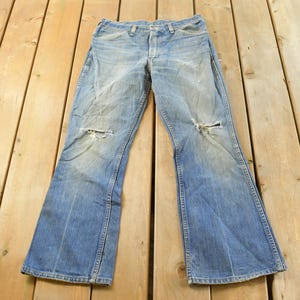 Puede incluir: Vaqueros de mezclilla azul claro con detalles desgastados, incluyendo roturas en las rodillas. Los vaqueros tienen un dise&ntilde;o de pierna acampanada y est&aacute;n sobre una superficie de madera.