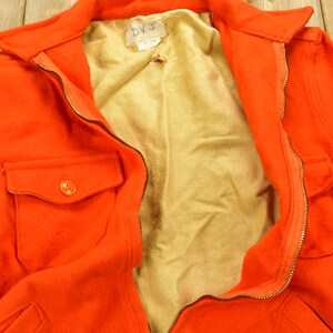 K&ouml;nnte beinhalten: Eine leuchtend orangefarbene Jacke mit Rei&szlig;verschluss und einer einzigen Knopftasche. Die Jacke ist ge&ouml;ffnet und zeigt ein hellbraunes Futter. Das Etikett auf der Jacke lautet "DVJ".