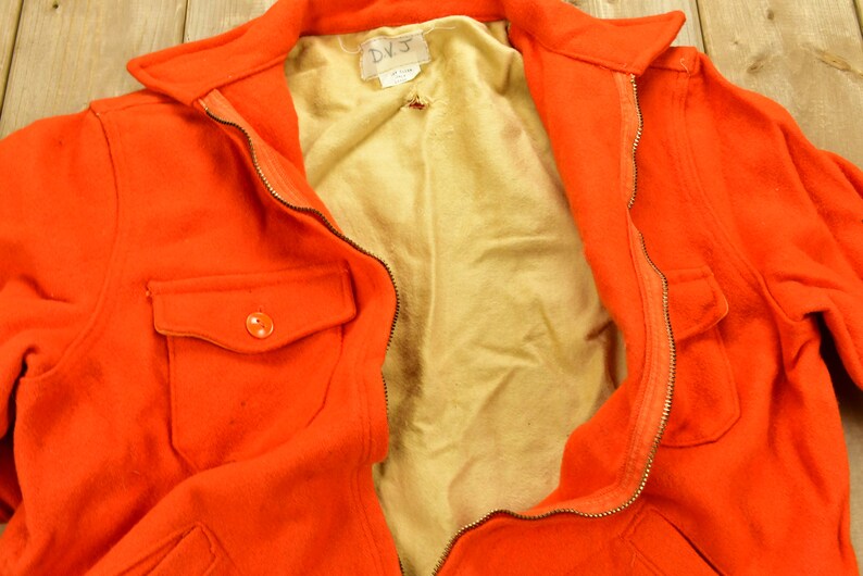 K&ouml;nnte beinhalten: Eine leuchtend orangefarbene Jacke mit Rei&szlig;verschluss und einer einzigen Knopftasche. Die Jacke ist ge&ouml;ffnet und zeigt ein hellbraunes Futter. Das Etikett auf der Jacke lautet "DVJ".