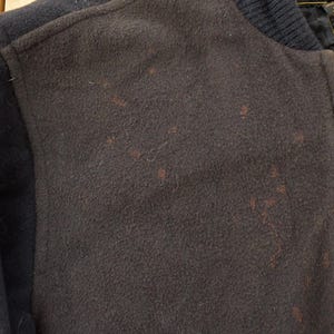 May include: A dark brown and black varsity jacket featuring a wool-like texture on the front panels and sleeves. The jacket includes a ribbed collar, a full-length front zipper, and a visible interior tag near the neckline with text in English.